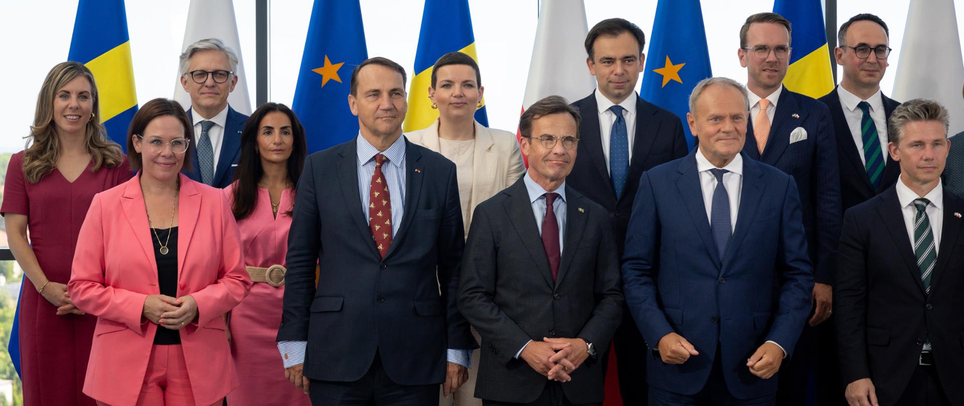 A group of twelve participants poses for a group photograph against a backdrop of the flags of Sweden, Poland and the European Union arranged in alternating order, and a glass wall offering a view of greenery. In the front row are Deputy Prime Minister Radosław Sikorski and the Prime Ministers of Sweden and Poland, Ulf Kristersson and Donald Tusk. Standing in the back row is Deputy Minister of Foreign Affairs Artur Harazim. The photograph is of an official and diplomatic nature.