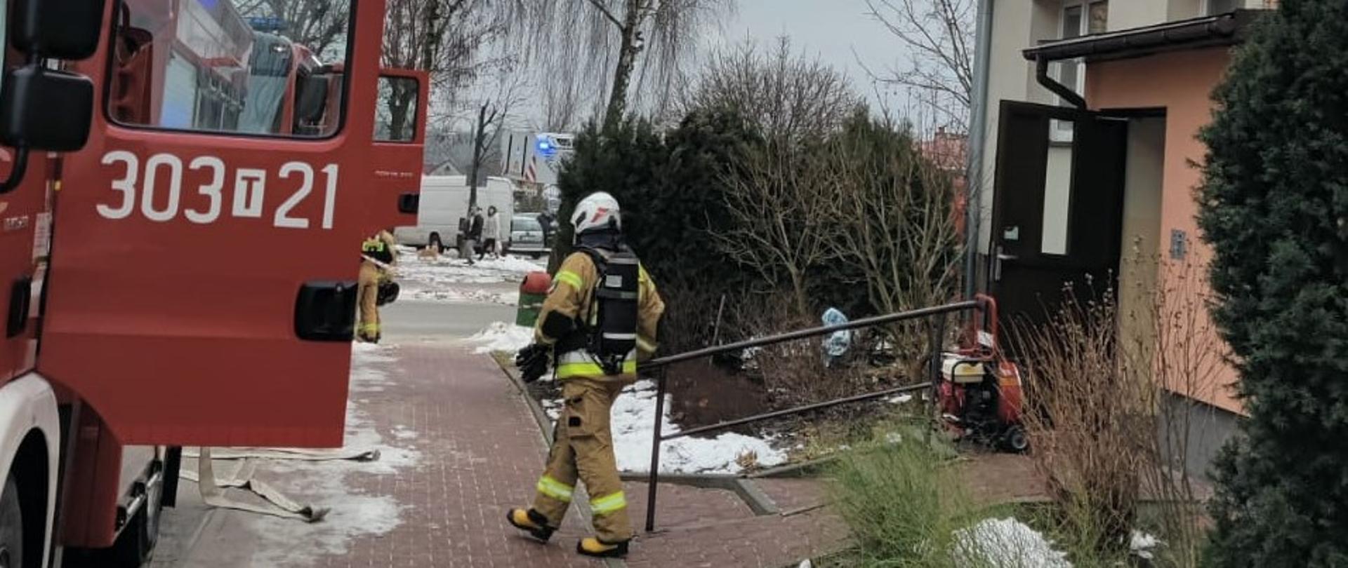 Na zdjęciu widzimy miejsce działań: z lewej strony samochód pożarniczy, z prawej budynek wielorodzinny, w którym wybuchł pożar. Ratownik idzie do samochodu, widać również przygotowany wentylator do oddymiania.