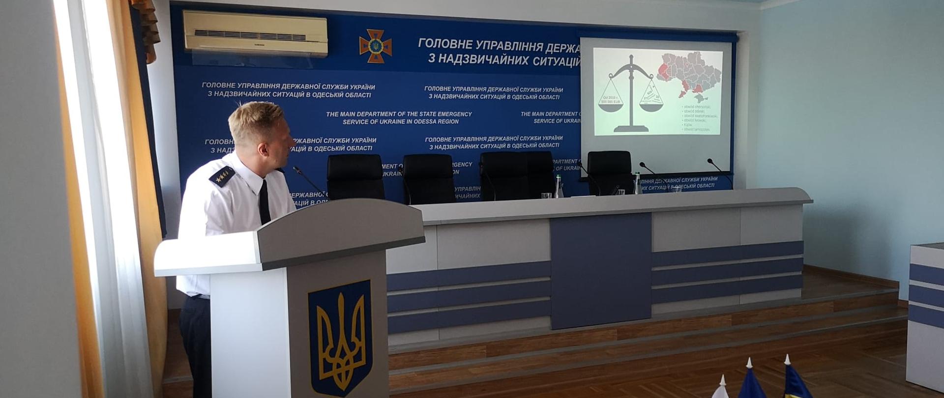 strażak w białej koszuli stoi za mównicą i prezentuje wyświetlony na dużym ekranie slajd z prezentacji;