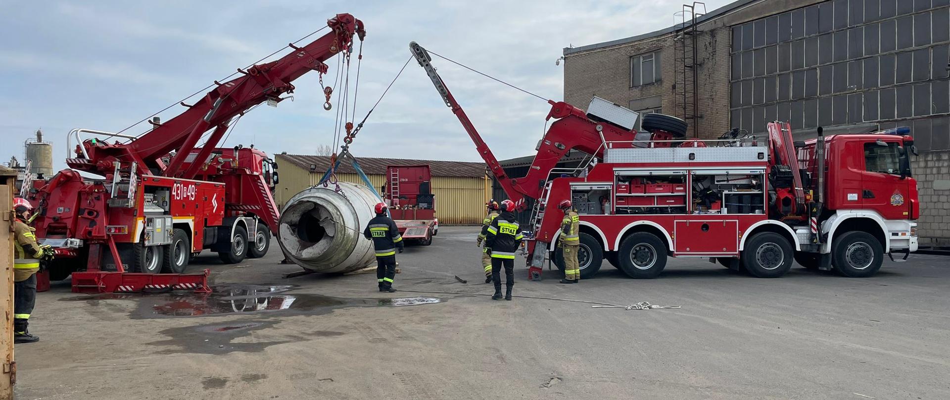 Zdjęcie przedstawia dwa ciężkie samochody ratownictwa drogowego, przy użyciu których operatorzy starają się podnieść beczkę wypełnioną betonem. Zdjęcie wykonano na terenie zakładu produkcyjnego.
