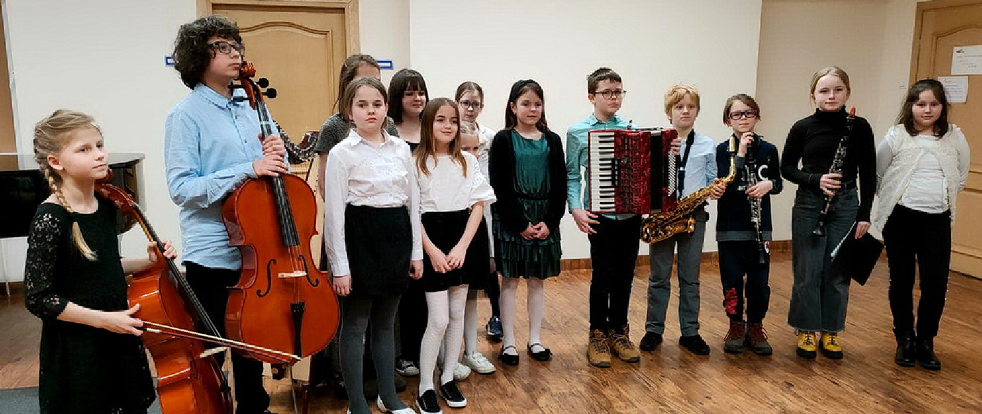 Na zdjęciu stoi grupa 13 dzieci, 9 dziewczynek i 4 chłopców. Dzieci stoją w auli szkolnej, twarzami są zwrócone w kierunku fotografującego, dwoje dzieci z lewej strony trzyma w rękach wiolonczele, dziecko z prawej strony trzymaj w rękach akordeon, kolejne dzieci saksofon i klarnety.