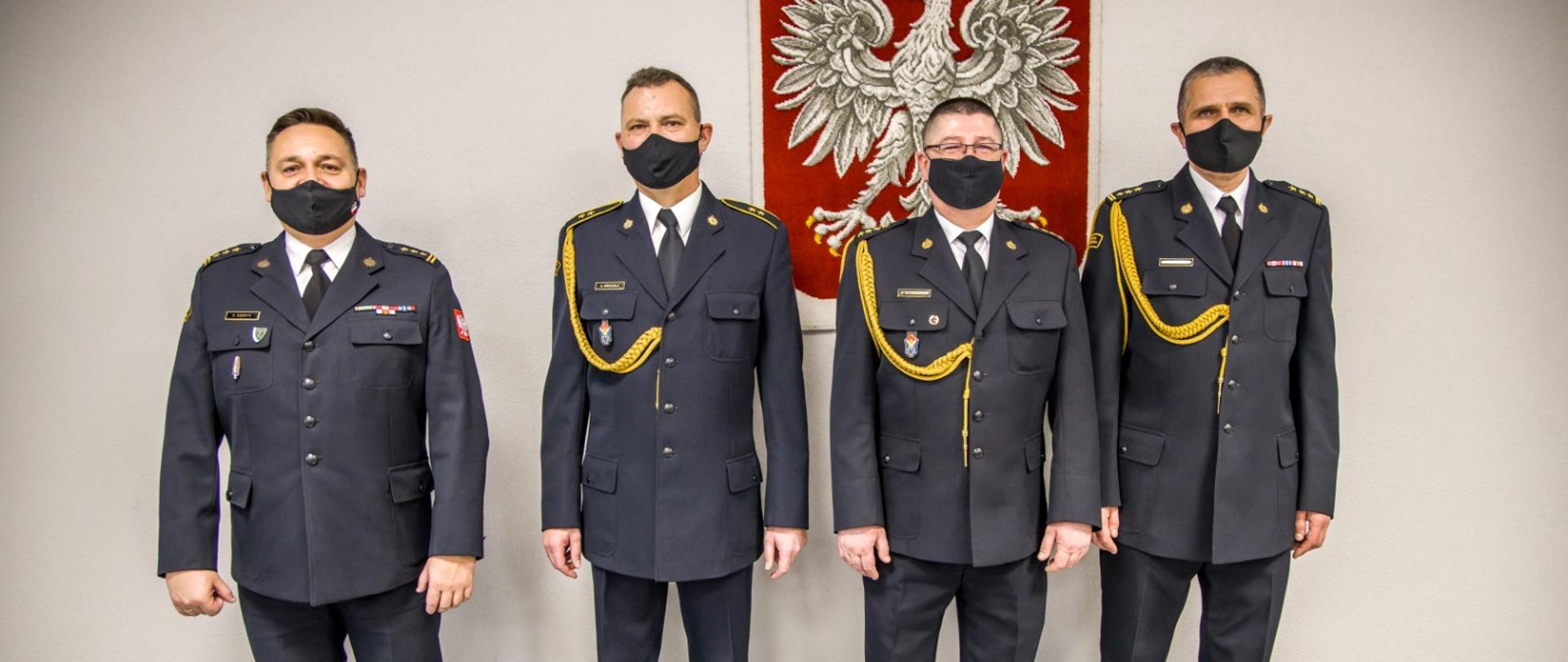 Zdjęcie funkcjonariuszy przechodzących na emeryturę w towarzystwie Opolskiego Komendanta Wojewódzkiego PSP w Opolu st. bryg. Krzysztofa Kędryka. Strażacy w mundurze galowym z założonymi na twarzy czarnymi maseczkami. W tle godło Polski powieszone na ścianie.