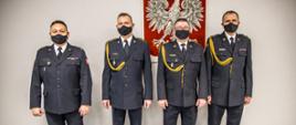 Zdjęcie funkcjonariuszy przechodzących na emeryturę w towarzystwie Opolskiego Komendanta Wojewódzkiego PSP w Opolu st. bryg. Krzysztofa Kędryka. Strażacy w mundurze galowym z założonymi na twarzy czarnymi maseczkami. W tle godło Polski powieszone na ścianie.