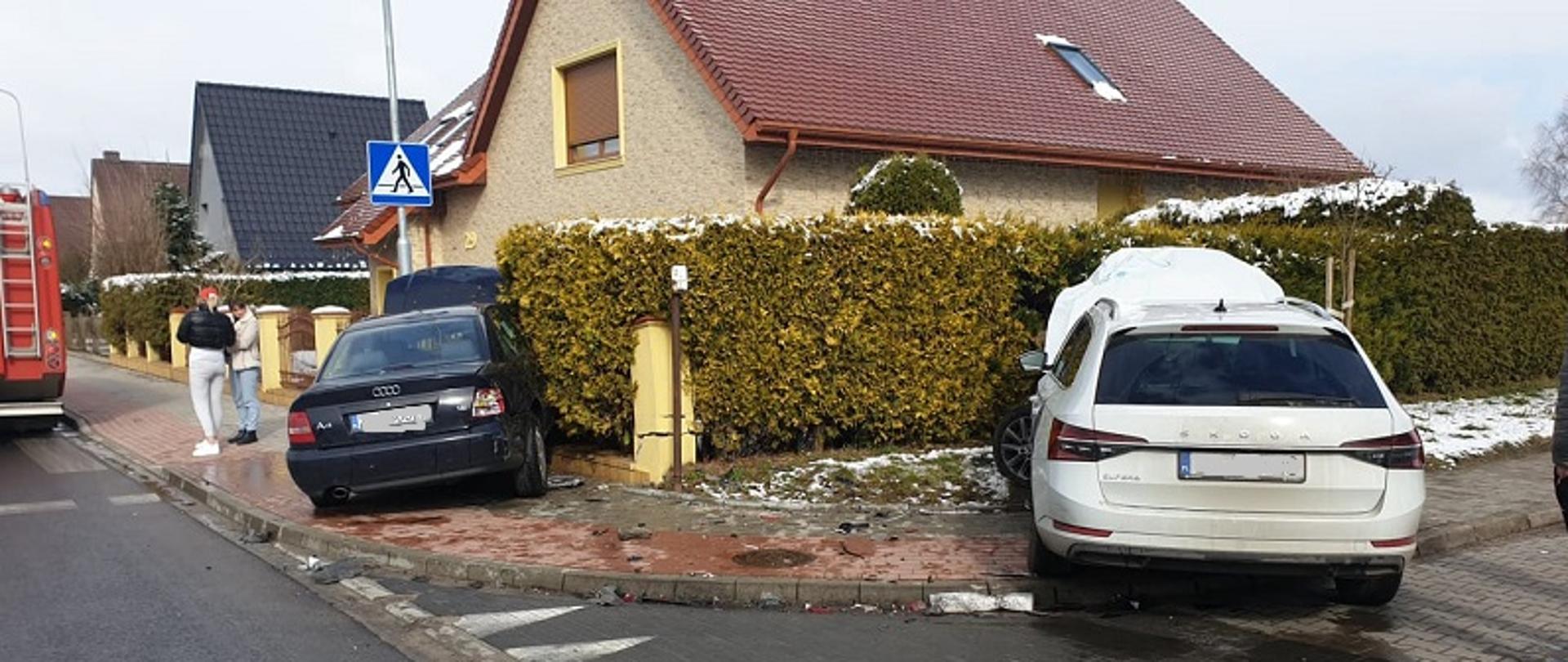 Zdjęcie przedstawia dwa pojazdy uczestniczące w zdarzeniu widoczne od tyłu. Ciemne Audi z otwartą maską znajduje się na chodniku przodem w żywopłocie posesji mieszkalnej. Biała Skoda znajduje się na chodniku po prawej stronie zdjęcia, posiada otwartą maskę przednią. W tle zdjęcia widoczne dachy domów budynków mieszkalnych.