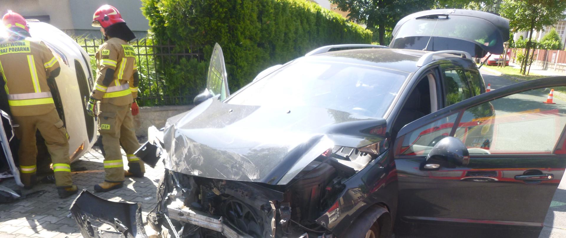 Samochód z rozbitym przodem i pootwieranymi drzwiami przednimi i klapą bagażnika, w tle strażacy