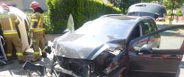 Samochód z rozbitym przodem i pootwieranymi drzwiami przednimi i klapą bagażnika, w tle strażacy