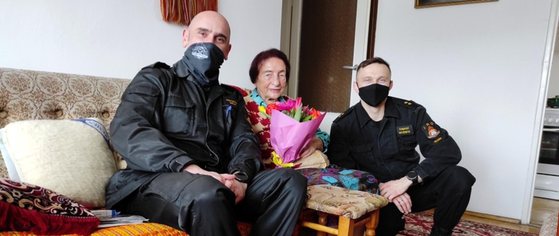 Na zdjęciu widać strażaków z wizytą u Seniorki - Pani Kazimiery, dzień po szczepieniu. Pani Kazimiera siedzi pomiędzy strażakami z otrzymanym bukietem kwiatów.