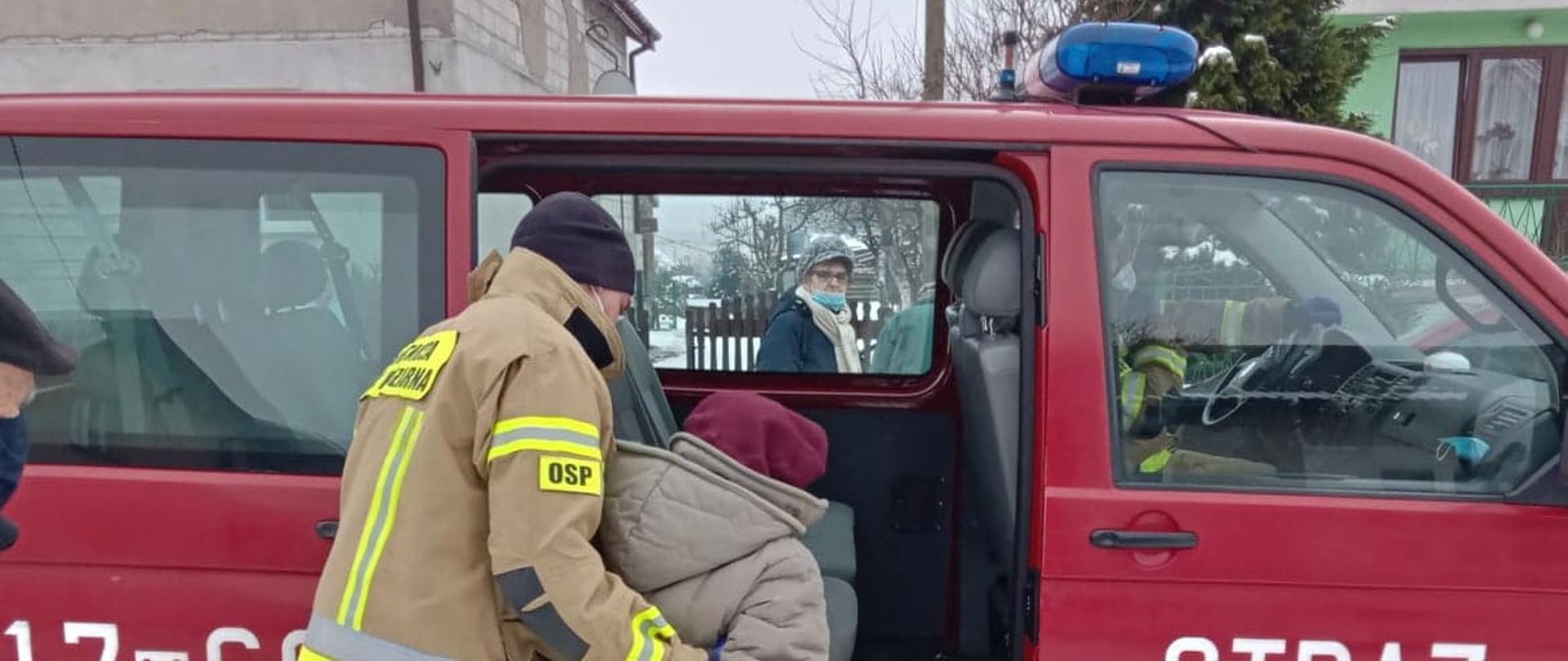 Strażak ochotniczej straży pożarnej ubrany w żółte ubranie specjalne pomaga wsiąść starszej kobiecie do czerwonego samochodu należącego do OSP. W tle budynki.

