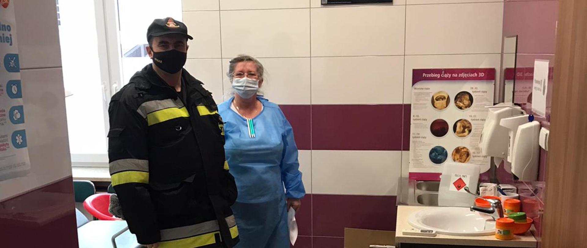 Na zdjęciu widać strażaka oraz pracowniczkę punktu szczepienia po przekazaniu środków ochrony osobistej. Zdjęcie wykonane w pomieszczeniu punktu szczepienia. W tle środki ochrony osobistej.