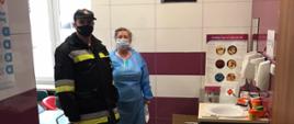 Na zdjęciu widać strażaka oraz pracowniczkę punktu szczepienia po przekazaniu środków ochrony osobistej. Zdjęcie wykonane w pomieszczeniu punktu szczepienia. W tle środki ochrony osobistej.