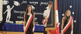 na zdjęciu troje uczniów, dwie dziewczynki ubrane na czarno i jeden chłopiec ubrany w czarne spodnie i białą koszulę, wszyscy mają na sobie szarfę biało-czerwoną, chłopiec stoi w środku i trzyma sztandar szkoły