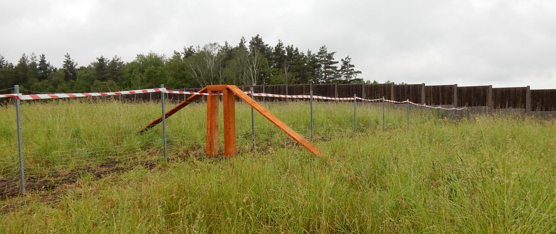 Na zdjęciu widać teren porośnięty wysoką, zieloną trawą. Przez środek biegnie prowizoryczne ogrodzenie z metalowych słupków i biało‑czerwonej taśmy. W centralnej części stoi drewniana konstrukcja w kształcie litery „A”, wspierająca taśmę. W tle znajduje się linia drzew oraz rząd pionowych, ciemnych stalowych grodzic. Niebo jest szare i pochmurne.
