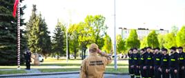 Na placu przed Jednostka Ratowniczo Gaśnicza nr 1 stoją strażacy w dwuszeregu na uroczystej zbiórce, ubrani w czarne ubrania koszarowe. Na środku stoi dowódca ubrany w piaskowy mundur. Na maszt uroczyście wciągana jest biało czerwona flaga przez poczet sztandarowy ubrany w mundury galowe.