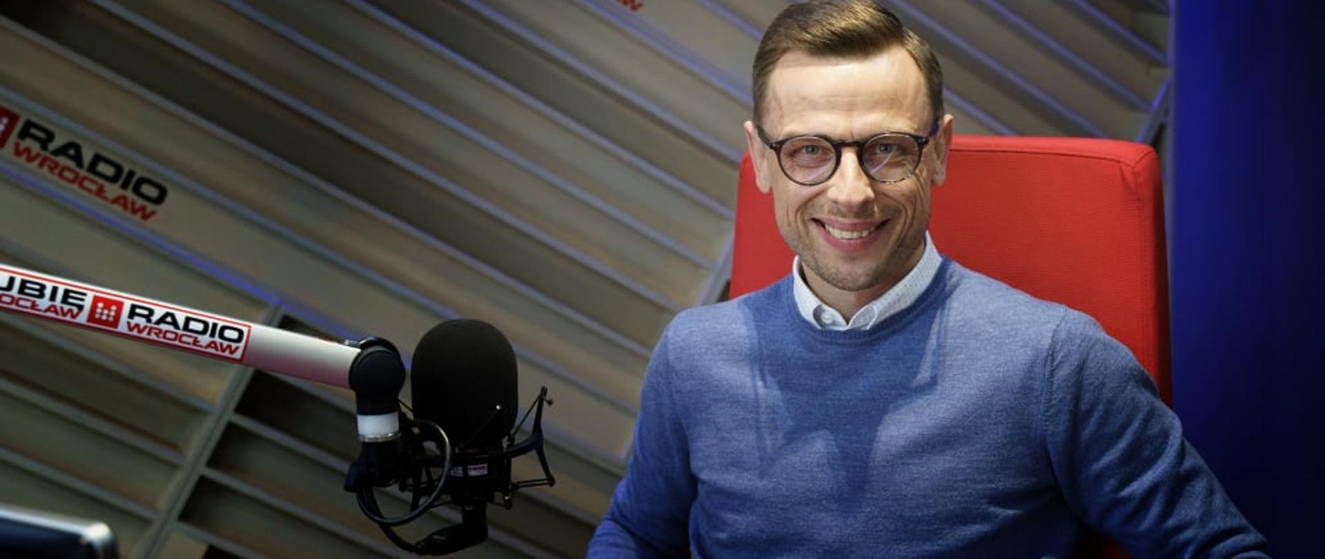 Dolnośląski Wojewódzki Inspektor Inspekcji Handlowej we Wrocławiu w studiu Radia Wrocław, mężczyzna ubrany jest w niebieski sweter, siedzi na czerwonym fotelu, przed nim na metalowym wysięgniku znajduje się mikrofon. 