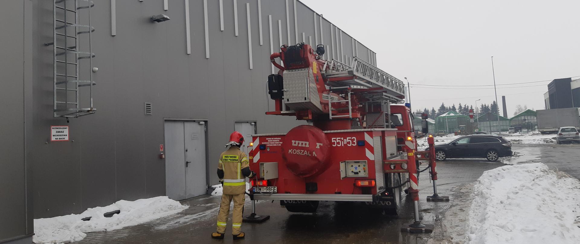 Na zdjęciu widać samochód pożarniczy - podnośnik hydrauliczny wraz ze strażakiem stojący przy budynku handlowym, w tle samochody osobowe na parkingu