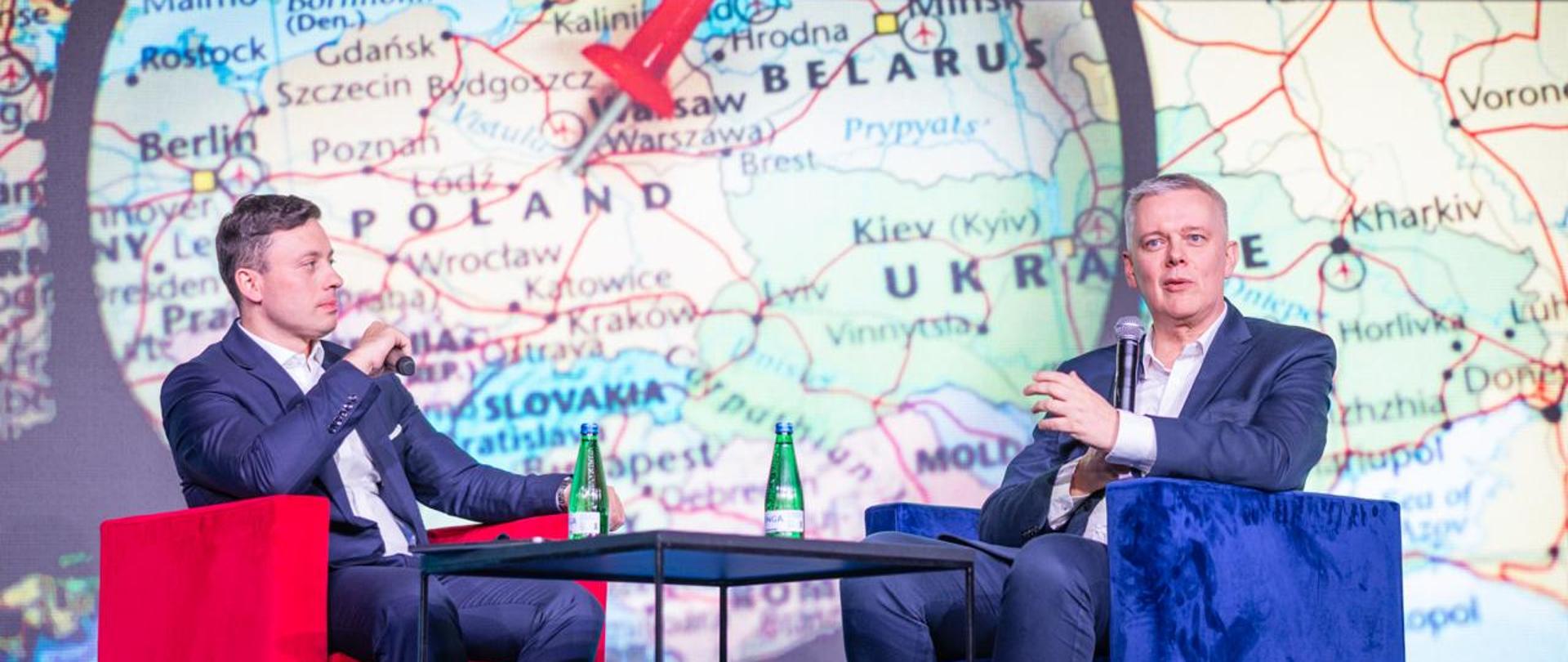 Po prawej stronie na niebieskim fotelu siedzi minister Tomasz Siemoniak, a po lewej stronie na czerwonym fotelu siedzi redaktor Piotr Witwicki. Na środku na czarnym stoliku stoją zielone butelki z wodą. Minister i dziennikarz trzymają w rękach mikrofony bezprzewodowe. W tle widać fragment mapy Europy pod lupą. Na mapie czerwoną pinezką oznaczona jest Warszawa.