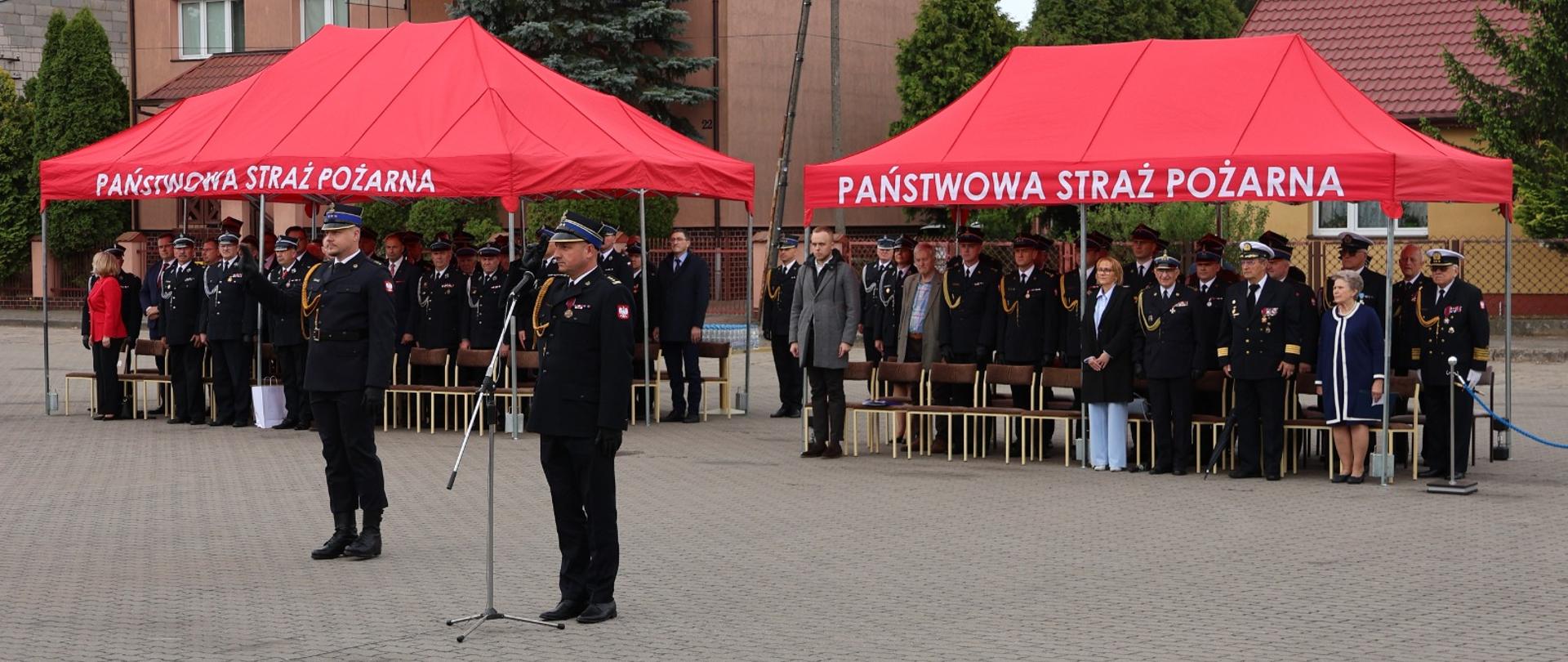 Do mikrofonu przemawia Zastępca Mazowieckiego Komendanta Wojewódzkiego PSP – st. bryg. Maciej Bieńczyk. W tle goście uroczystości pod czerwonymi namiotami z napisem „PAŃSTWOWA STRAŻ POŻARNA”.
