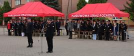 Do mikrofonu przemawia Zastępca Mazowieckiego Komendanta Wojewódzkiego PSP – st. bryg. Maciej Bieńczyk. W tle goście uroczystości pod czerwonymi namiotami z napisem „PAŃSTWOWA STRAŻ POŻARNA”.