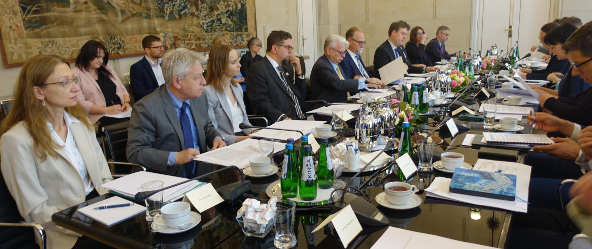 Grupa osób siedzi przy stole, na którym są mikrofony i notatki. 