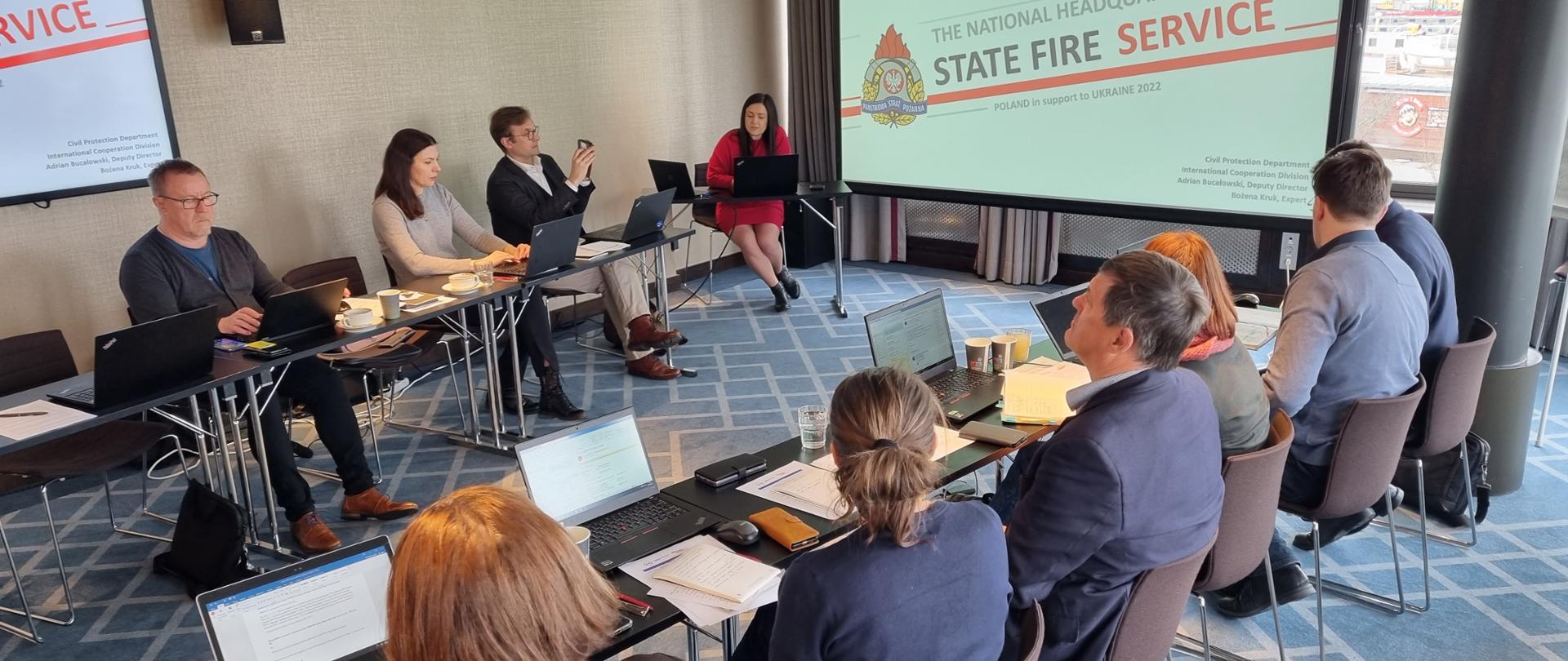 dziesięć osób siedzący przy dwóch stołach w trakcie spotkania roboczego. Na stołach widoczne laptopy oraz notatniki. Na widocznym w tle monitorze wyświetlana jest prezentacja