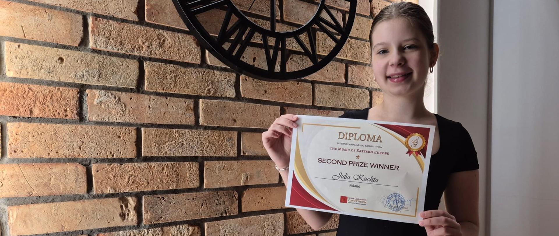 Uczennica Julka stoi na tle ceglanej ściany, trzyma dyplom. Na ścianie zegar.
