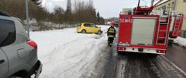 Na zdjęciu widoczne tyły dwóch pojazdów pożarniczych z przechodzącym strażakiem. Z lewej strony widoczny szary fragment tylnej bocznej strony samochodu na ośnieżonym poboczu ulicy, w głębi widoczna prawa boczna strona żółtego samochodu z uszkodzonym przodem stojącego na śniegu. Widoczne błoto pośniegowe na jezdni.