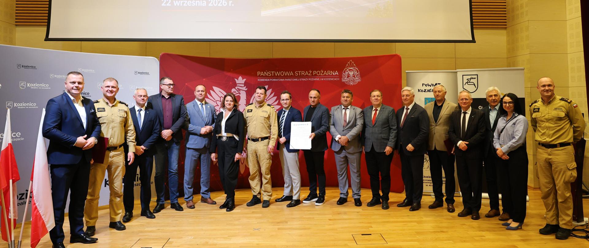 Grupa uczestników uroczystości stoi przed ekranem z prezentacją dotyczącą podpisania umowy na budowę nowej Komendy PSP w Kozienicach.