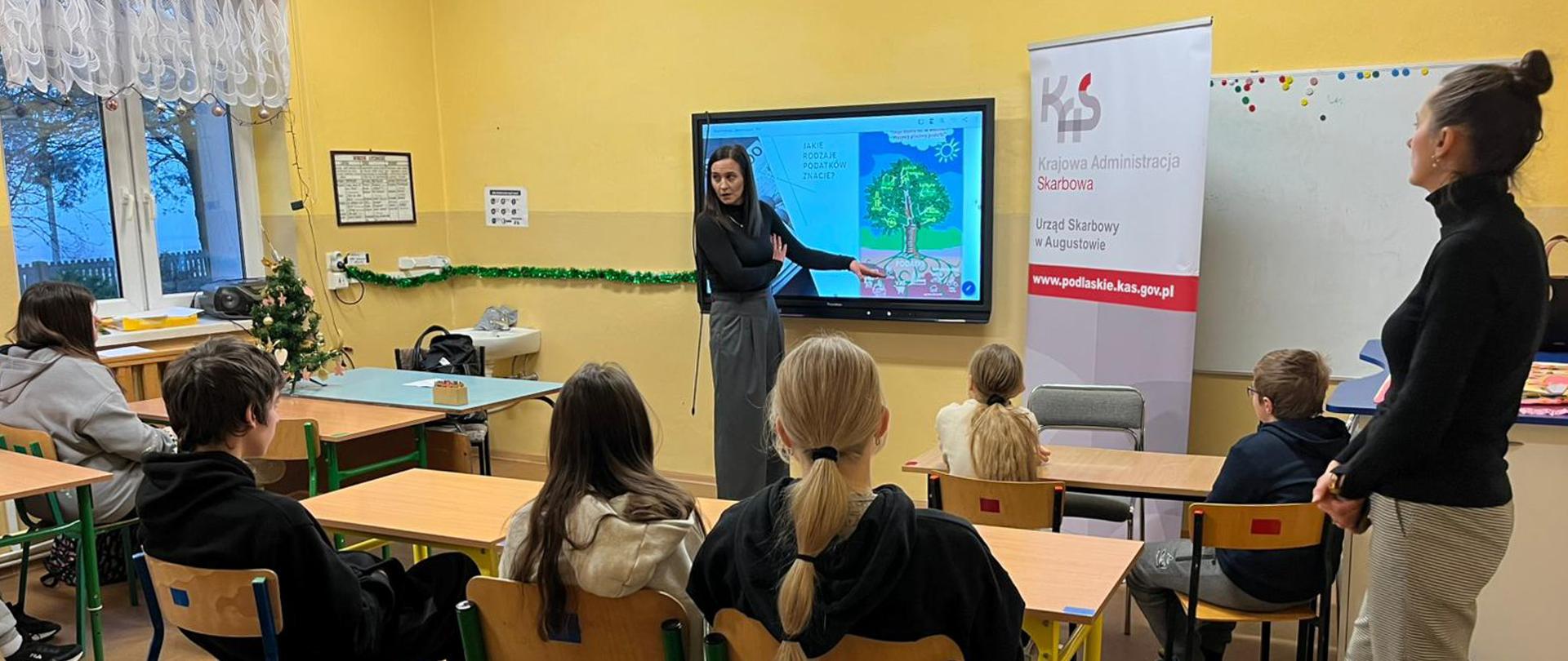 Pracownik US w Augustowie prowadzi lekcję o tematyce podatkowej w szkole podstawowej, w tle prezentacja z drzewem podatkowym: jakie rodzaje podatków znacie?, dzieci w ławkach, roll up Krajowej Administracji Skarbowej