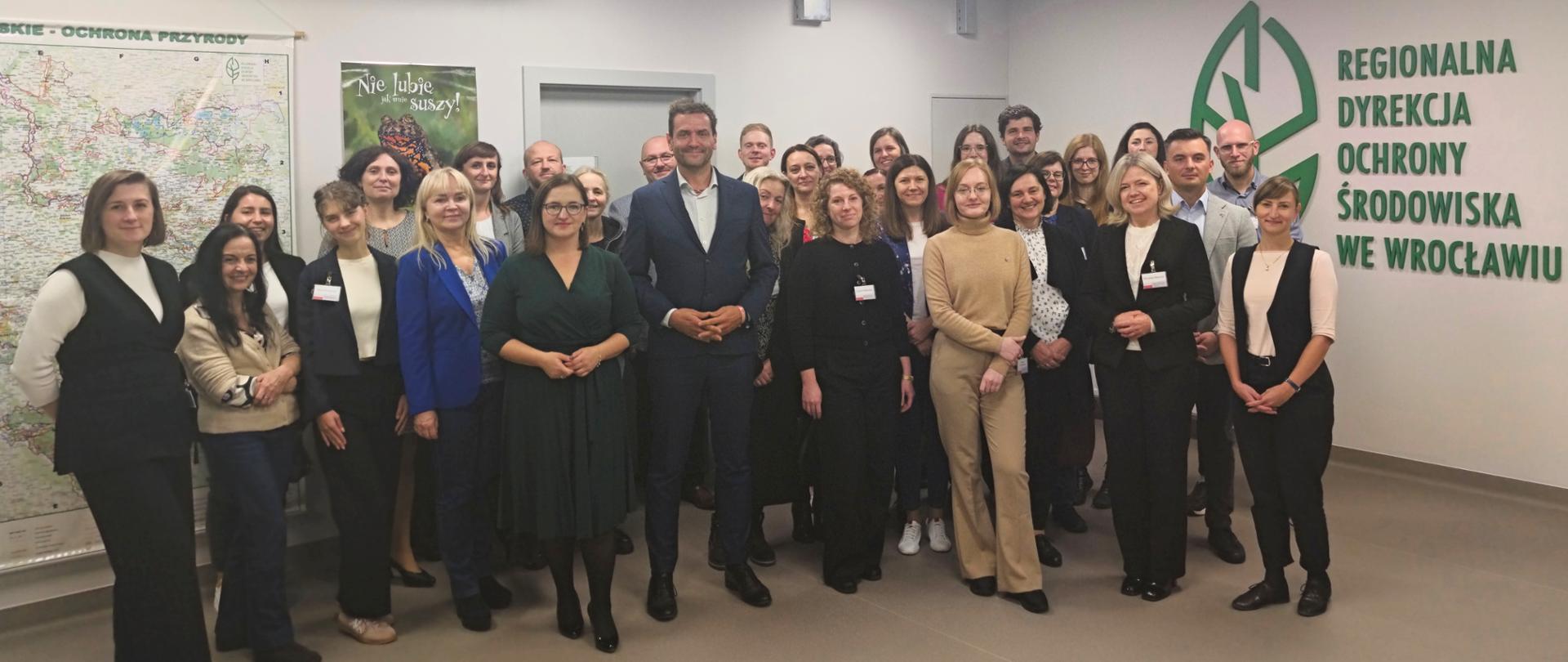 W tle przestrzeń biurowa, korytarz. Duża grupa elegancko ubranych, uśmiechniętych osób stoi obok siebie. Za ich placami logotyp Regionalnej Dyrekcji Ochrony Środowiska we Wrocławiu (zielony liść). 