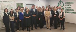 W tle przestrzeń biurowa, korytarz. Duża grupa elegancko ubranych, uśmiechniętych osób stoi obok siebie. Za ich placami logotyp Regionalnej Dyrekcji Ochrony Środowiska we Wrocławiu (zielony liść). 