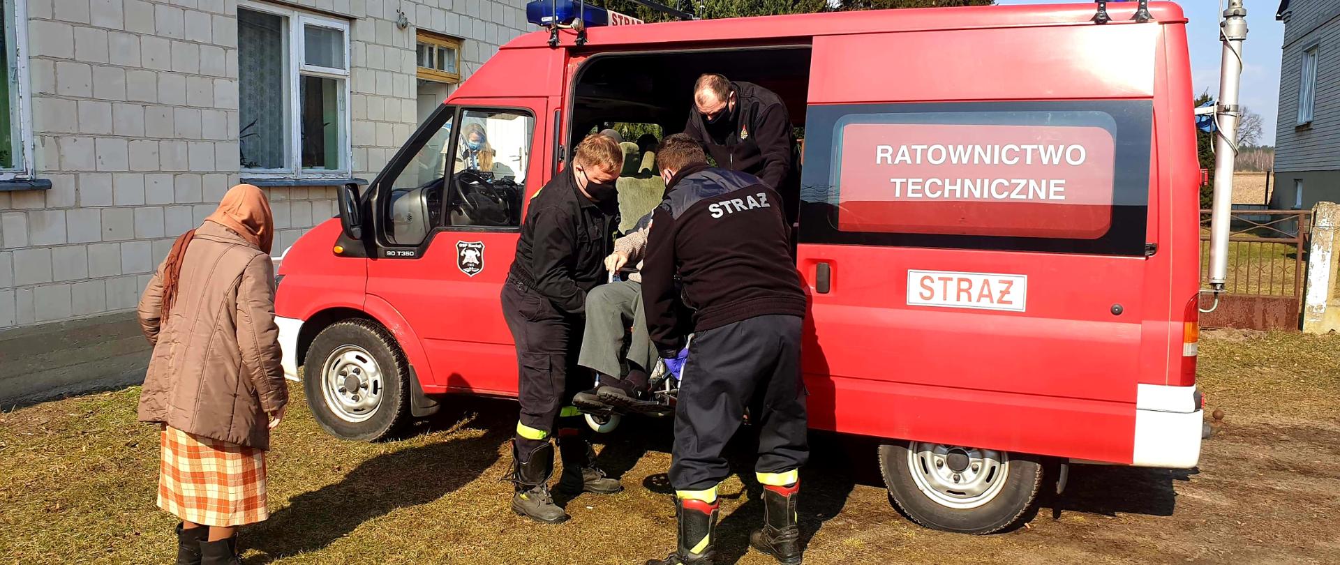Trzech strażaków OSP wyjmują z samochodu pożarniczego mężczyznę siedzącego na wózku inwalidzkim. Zdarzeniu przygląda się starsza osoba. Z lewej budynek mieszkalny z białej cegły.