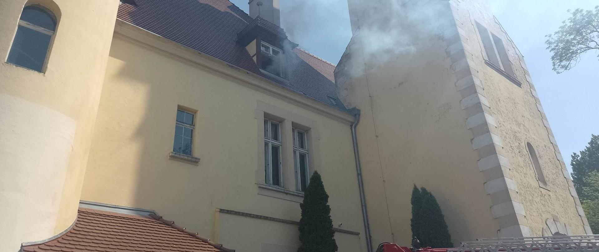 Zdjęcie przedstawia dym wydobywający się z otwartego okna na ostatniej kondygnacji budynku.