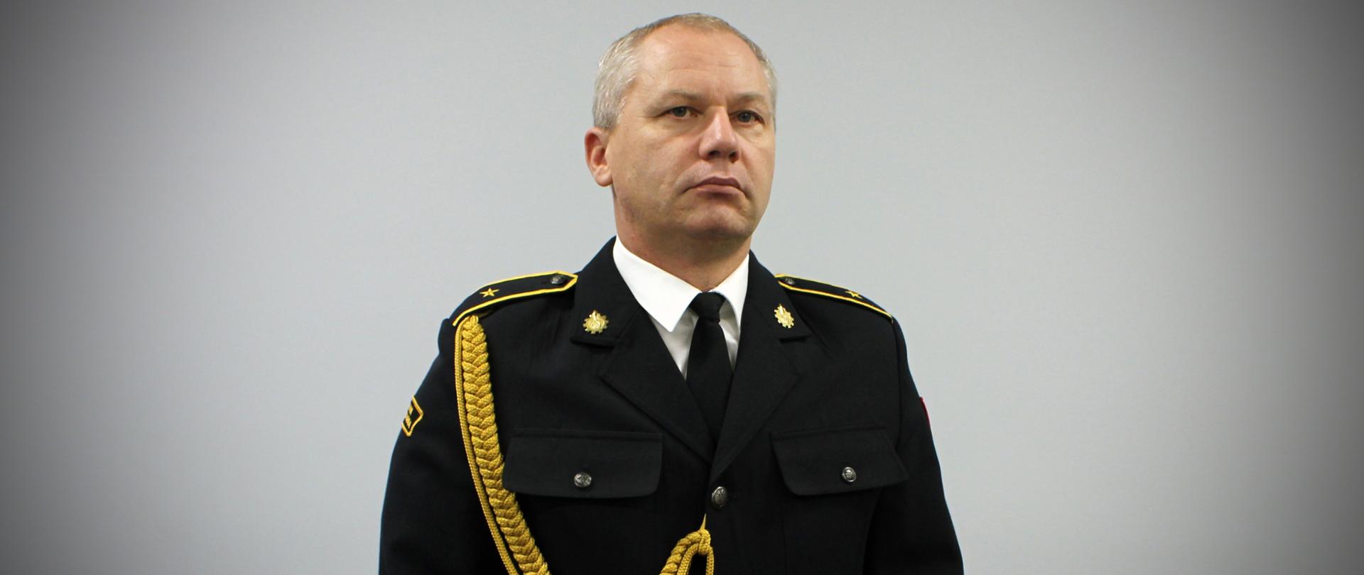 Na zdjęciu widać mężczyznę w eleganckim, galowym mundurze strażackim. Stoi na wprost obiektywu, w wyprostowanej, oficjalnej pozie. Mundur jest ciemny, z pagonami na ramionach, ozdobnym złotym sznurem na prawej stronie oraz kilkoma odznaczeniami i naszywkami. Na piersi znajduje się plakietka z nazwiskiem oraz niewielkie przypinki i barety świadczące o ukończonych szkoleniach lub wyróżnieniach. Mężczyzna ma krótkie, schludnie uczesane włosy i delikatny uśmiech. Tło jest jednolite, jasnoszare, dzięki czemu cała uwaga skupia się na postaci w mundurze