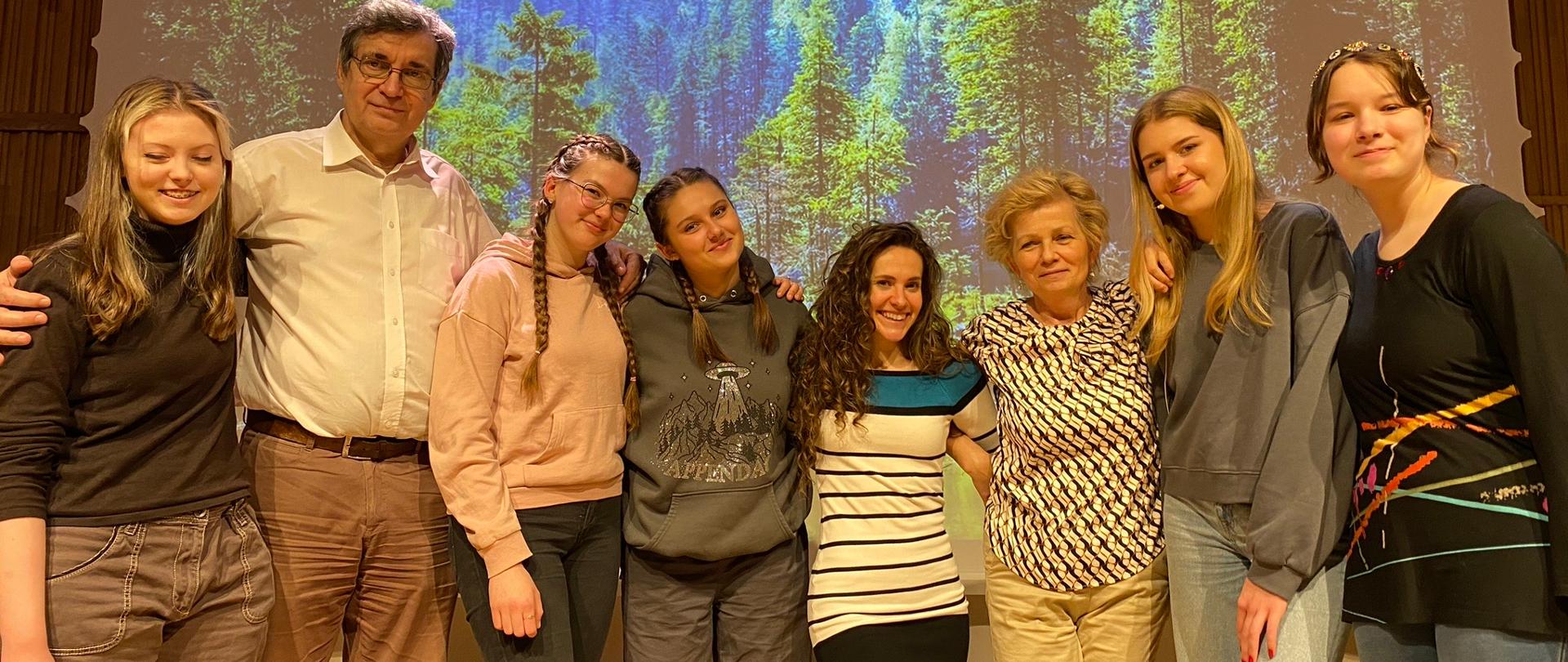 Zdjęcie. Na estradzie stoi grupa uczestników warsztatów musicalowych. W tle na dużym ekranie wyświetlony jest obraz lasu w górach.