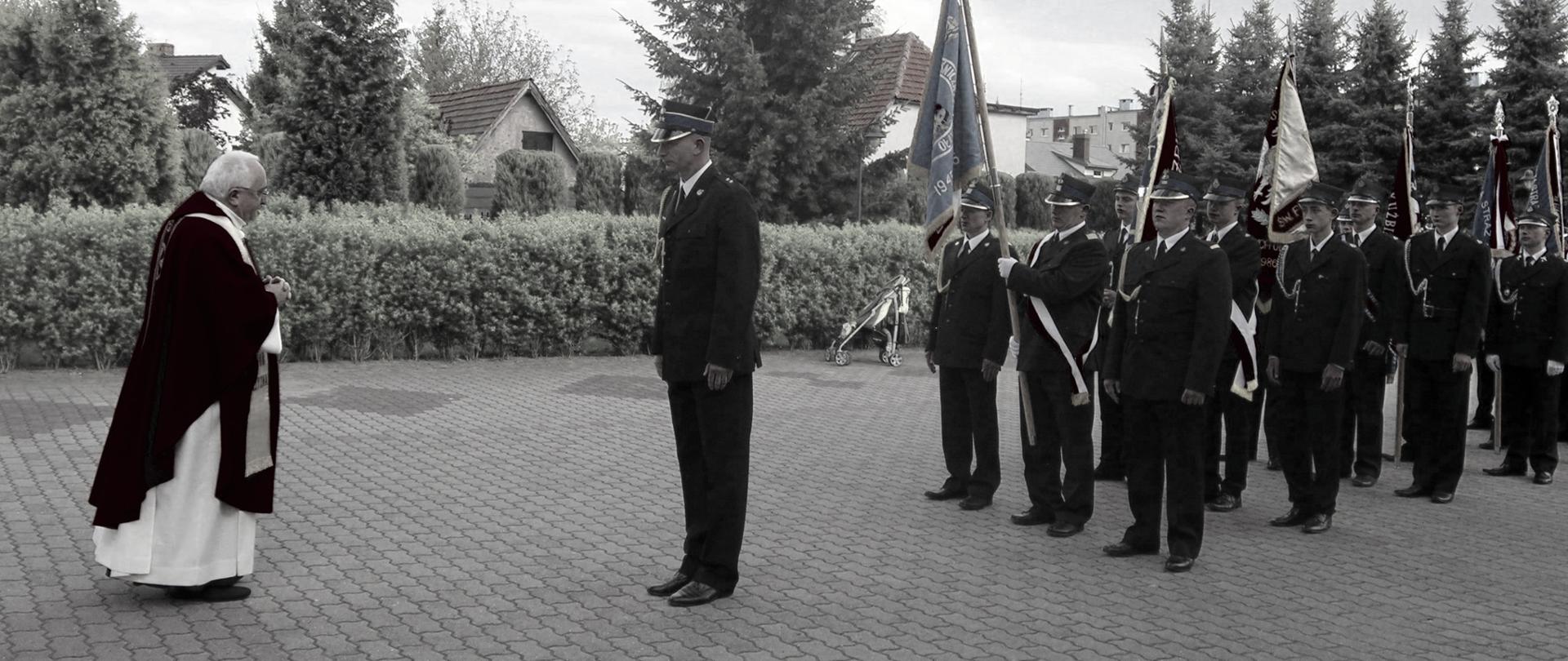 Ksiądz Stanisław Bijak wita procesje strażaków, na czele kpt. Bartłomiej Marcinów. Za nim poczty sztandarowe.