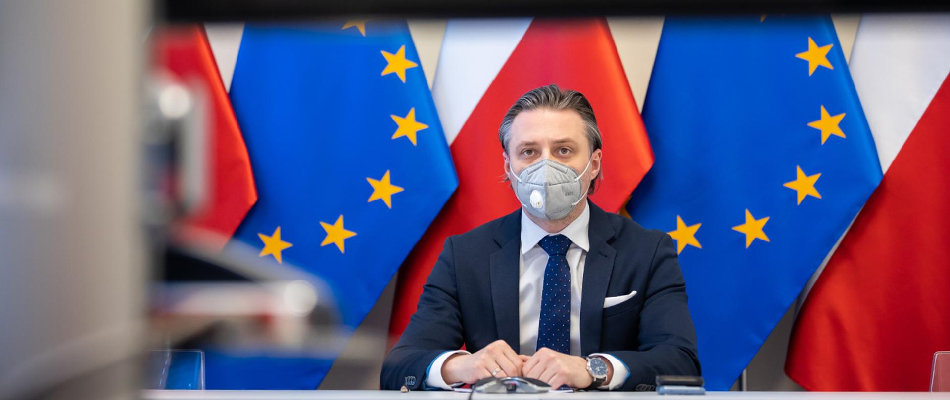 Na zdjęciu widać wiceministra Bartosza Grodeckiego wpatrzonego w ekran siedzącego na tle flag Polski i UE
