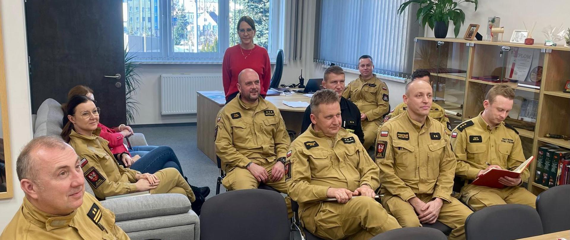 W jasnym pomieszczeniu odbywa się szkolenie. Kilku funkcjonariuszy w beżowych mundurach Państwowej Straży Pożarnej siedzi w dwóch rzędach. W tle widać regał z dokumentami i pamiątkami, a także duże okno z pionowymi żaluzjami. Atmosfera wygląda na spokojną i merytoryczną – część osób słucha, jedna osoba robi notatki, a dwie osoby w cywilnych ubraniach siedzą z boku, również uczestnicząc w spotkaniu.