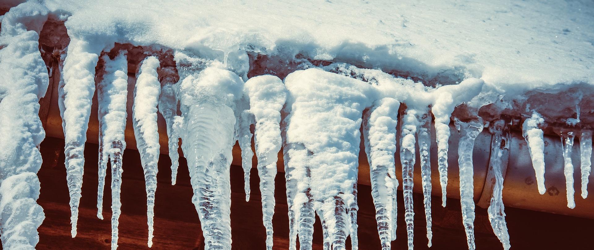 Zdjęcie przedstawia zalegający śnieg na dachu oraz zwisające sople lodowe