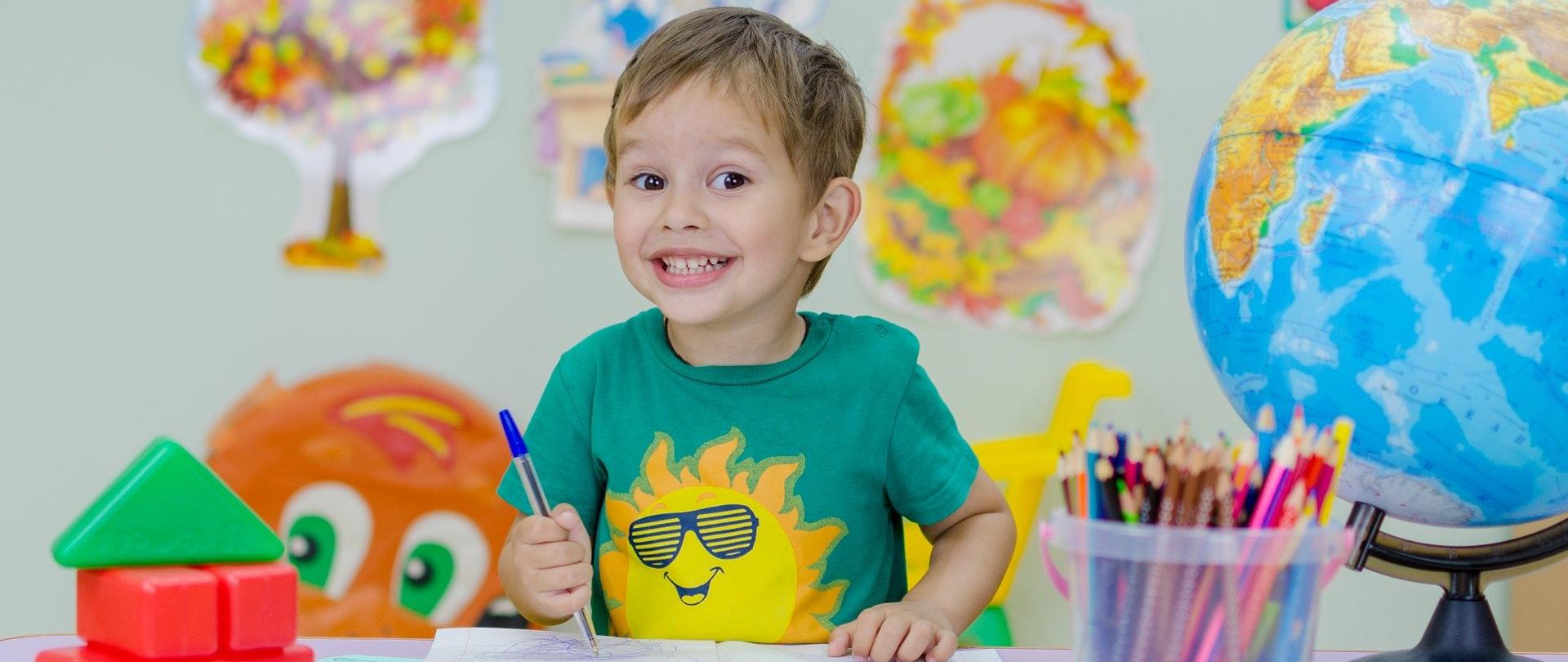 W centralnej części zdjęcia znajduje się uśmiechnięty chłopiec, siedzący za stołem. Maluje rysunek długopisem. Na stole, przy którym siedzi znajduje się przybory szkolne oraz - po prawej stronie - duży globus. W tle kolorowa ściana a na niej liczne naklejki edukacyjne. 