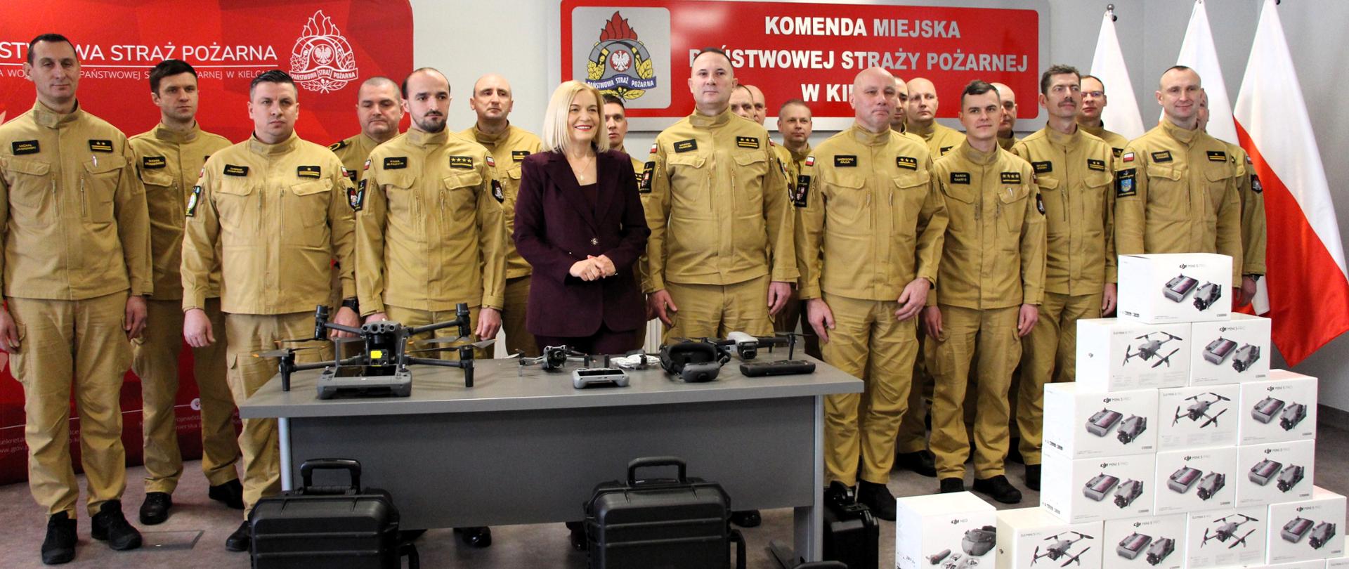 Na zdjęciu widać grupę strażaków Państwowej Straży Pożarnej stojących w sali konferencyjnej przy stole z dronami i walizkami sprzętu, na tle banerów PSP i flag Polski. Pośrodku stoi pani Marszałek województwa świętokrzyskiego oraz Świętokrzyski Komendant Wojewódzki PSP.