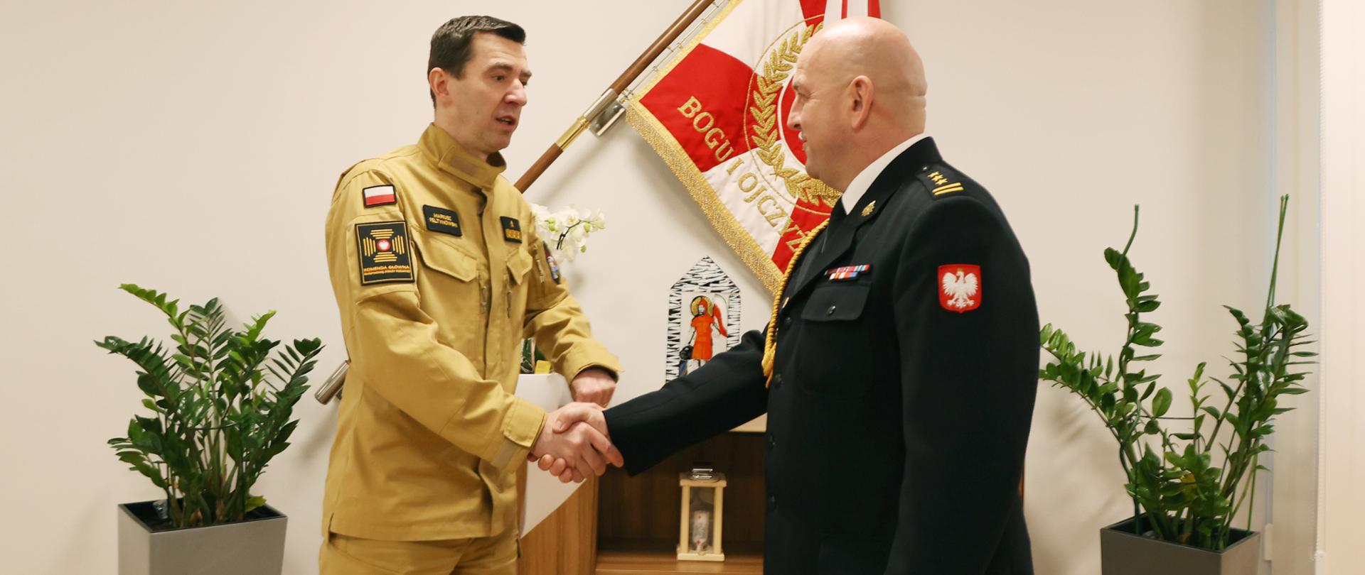 Dwaj strażacy., jeden w mundurze piaskowym, drugi w ciemnym galowym, ściskają sobie dłonie na tle sztandaru Komendy Głównej Państwowej Straży Pożarnej