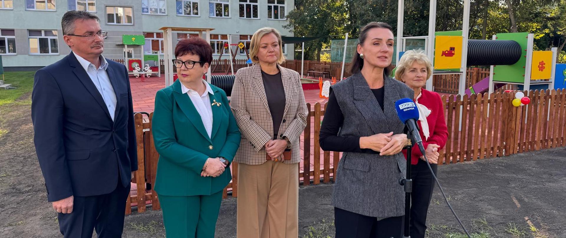 kilka osób stoi przed placem zabaw, jedna z kobiet przemawia