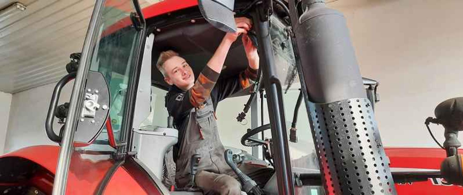 W ciągnichu rolniczym siedzi chłopak. Chłopak ma na sobie stroj roboczy, poprawia lusterko w ciągniku.