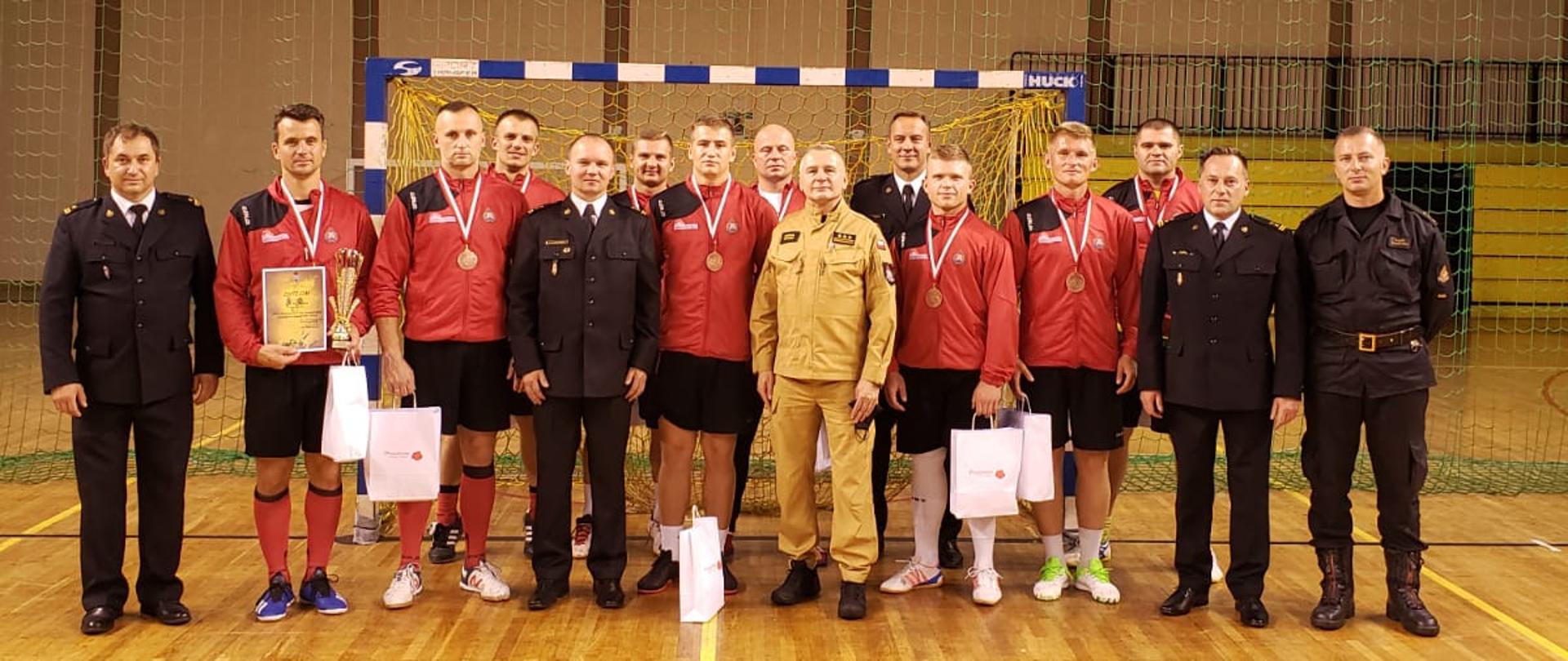 Zdjęcie przedstawia reprezentację Komendy Miejskiej Państwowej Straży Pożarnej w Radomiu podczas rozgrywek finałowych strażaków w piłkę nożną. Strażacy ustawieni są na tle bramki hali sportowej. Reprezentanci ubrani są w czerwono - czarne stroje piłkarskie (koszulki, spodenki). W śród nich znajdują się również zastępca mazowieckiego komendanta wojewódzkiego starszy brygadier Dariusz Sztumski, komendant miejski Państwowej Straży Pożarnej w Radomiu brygadier Paweł Fliszkiewicz wraz ze swoim zastępcą brygadierem Bartoszem Musiałem. Ponadto na zdjęciu widać jeszcze przedstawicieli organizatora zawodów funkcjonariuszy komendy powiatowej Państwowej Straży Pożarnej w Pruszkowie oraz ośrodka szkolenia w Warszawie. 