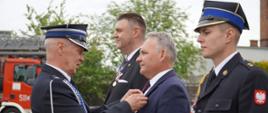 Podczas oficjalnych obchodów dnia strażaka jeden z druhów przypina odznaczenie mężczyźnie w garniturze. Obok stoją strażacy w mundurach galowych, a w tle widoczny jest samochód pożarniczy.
