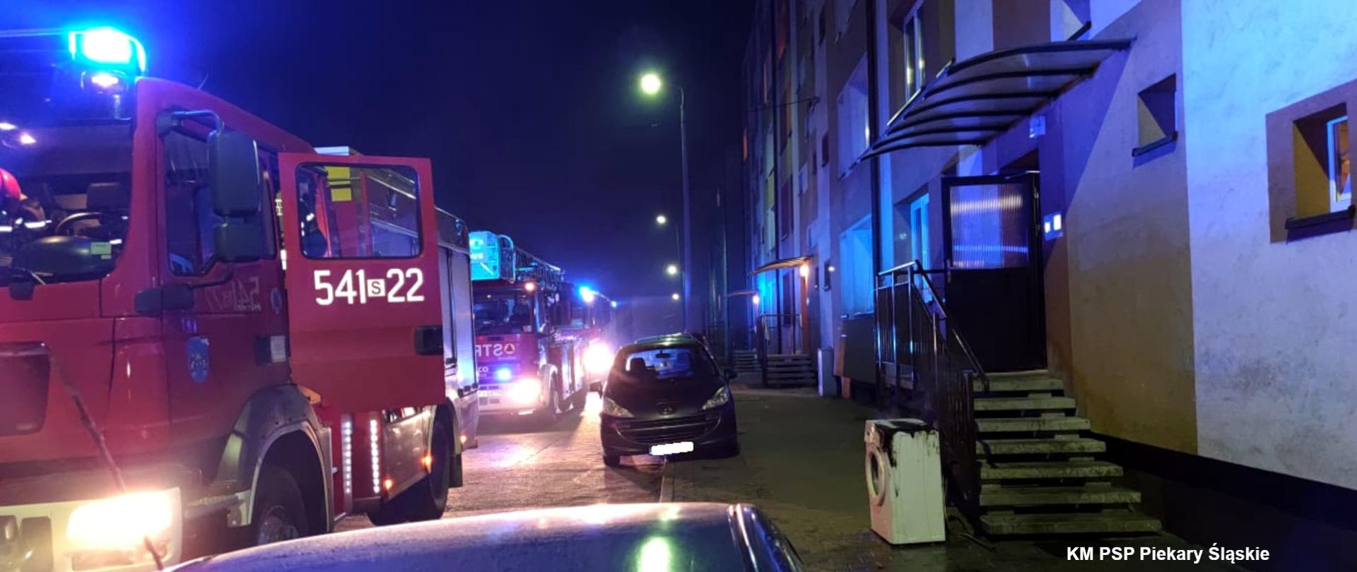 Zdjęcie przedstawia średni samochód ratowniczo - gaśniczy oraz samochód specjalny drabina mechaniczna z włączonymi światłami ostrzegawczymi stojące przed budynkiem mieszkalnym.