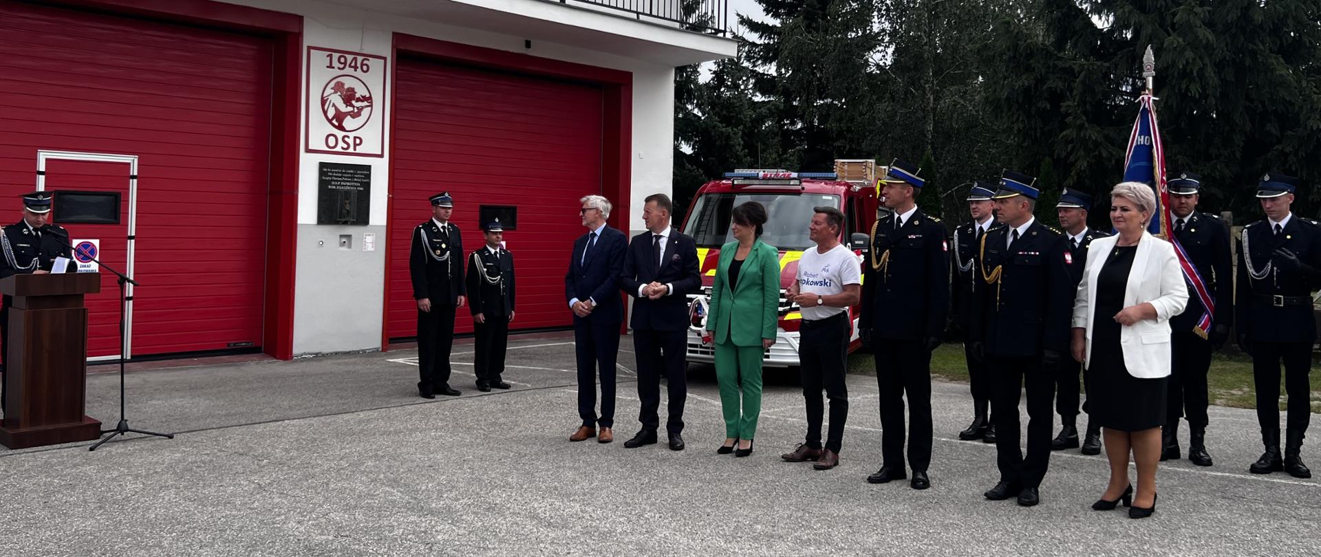 Zdjęcie przedstawia zaproszonych gości na uroczystość przekazania nowego samochodu strażackiego dla druhów z OSP Paprotnia. Z tyłu widać nowy pojazd oraz druhów stojących w dwuszeregu 