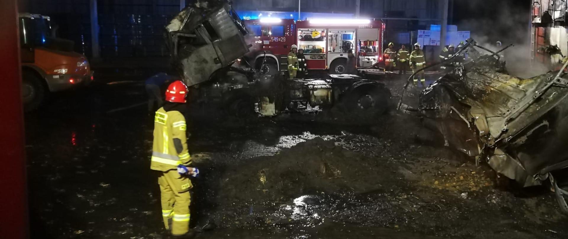 Zdjęcie przedstawia działania gaśnicze podczas pożaru samochodów ciężarowych. Na zdjęciu widzimy wraki pojazdów na tle wozu bojowego biorącego udział w działaniach oraz strażaka podczas oględzin miejsca zdarzenia. Zdjęcie wykonane w porze nocnej na placu składowym.