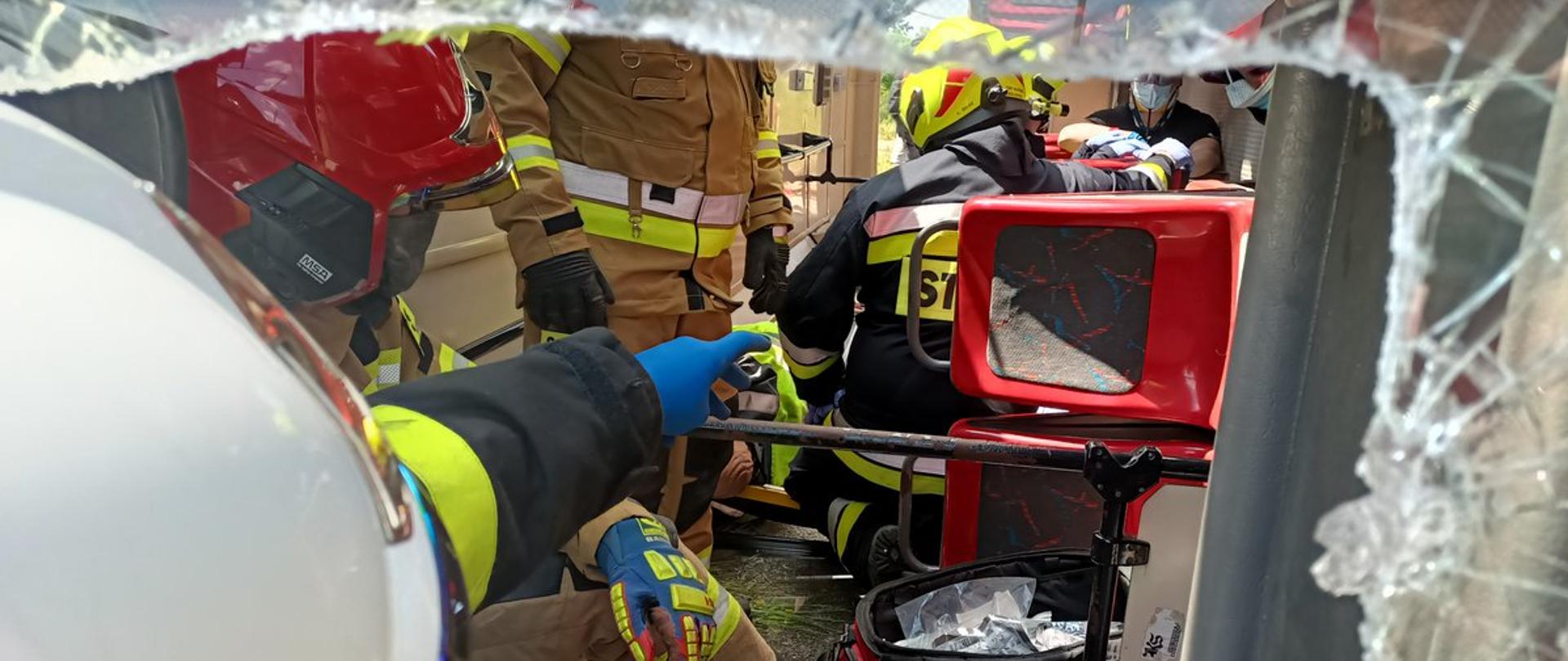 Zdjęcie przedstawia wnętrze do wywróconego autobusu. Zdjęcie wykonane przez rozbitą szybę pojazdu. W środku autobusu strażacy prowadzą działania ratownicze. 