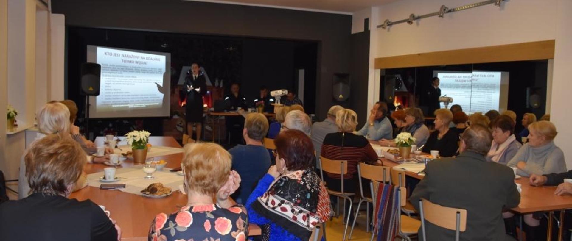 Zdjęcie wykonane na sali Gminnym Centrum Kultury i Biblioteki w Wolanowie. Na zdjęciu widać mieszkańców gminy Wolanów uczestniczących w pogadance tematycznej „czujka na straży twojego bezpieczeństwa”. Zgromadzone na sali osoby siedzą na krzesłach ustawionych przy stołach. W tle widać rzutnik multimedialny oraz ekran na którym funkcjonariuszka z Komendy Miejskiej Państwowej Straży Pożarnej w Radomiu wyświetla prezentacje. 